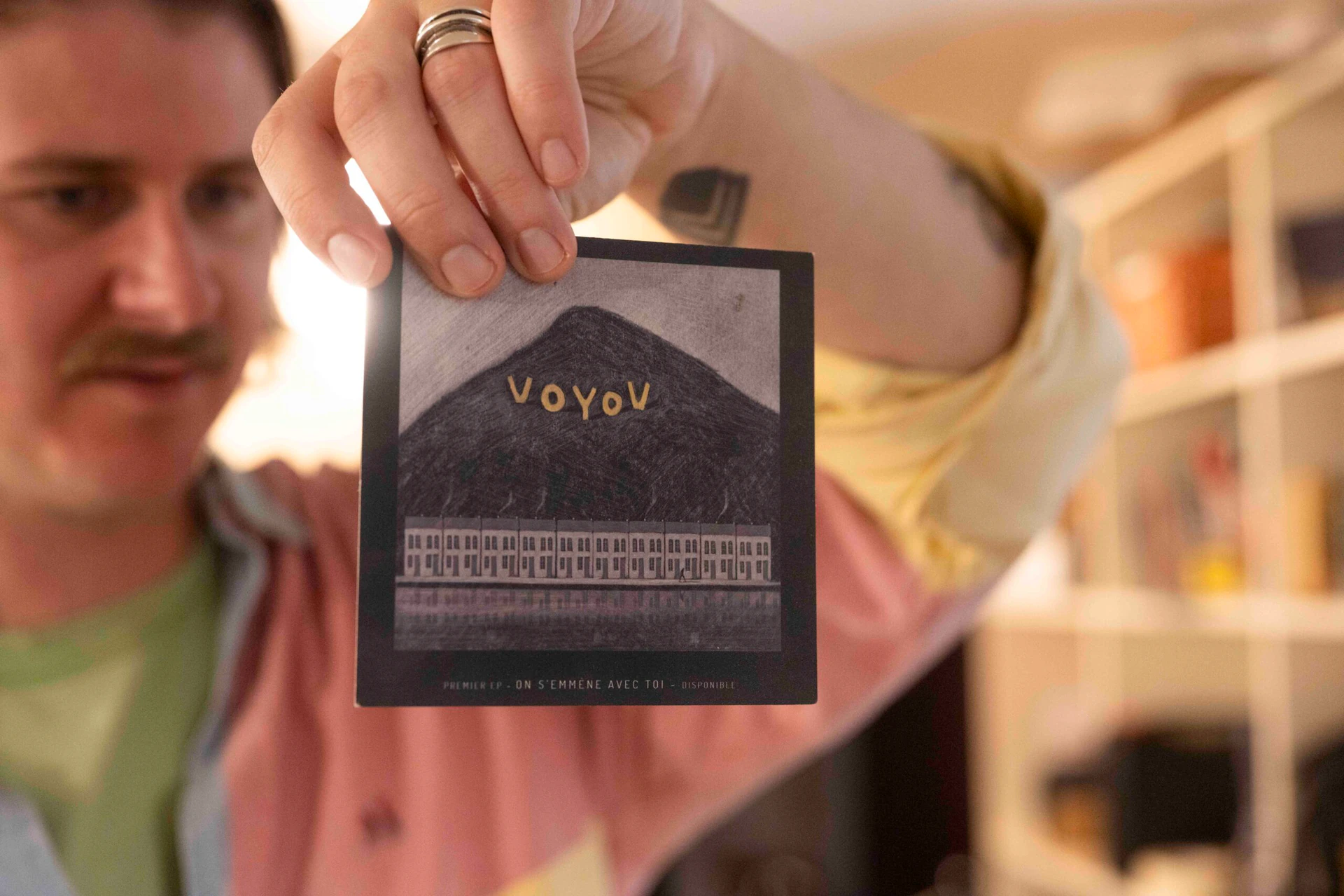 Main tenant la pPochette du premier EP de Voyou, « On s'emmène avec toi », dessin d'une montagne surmontant des immeubles, avec Thibaud Vanhooland flou en arrière-plan